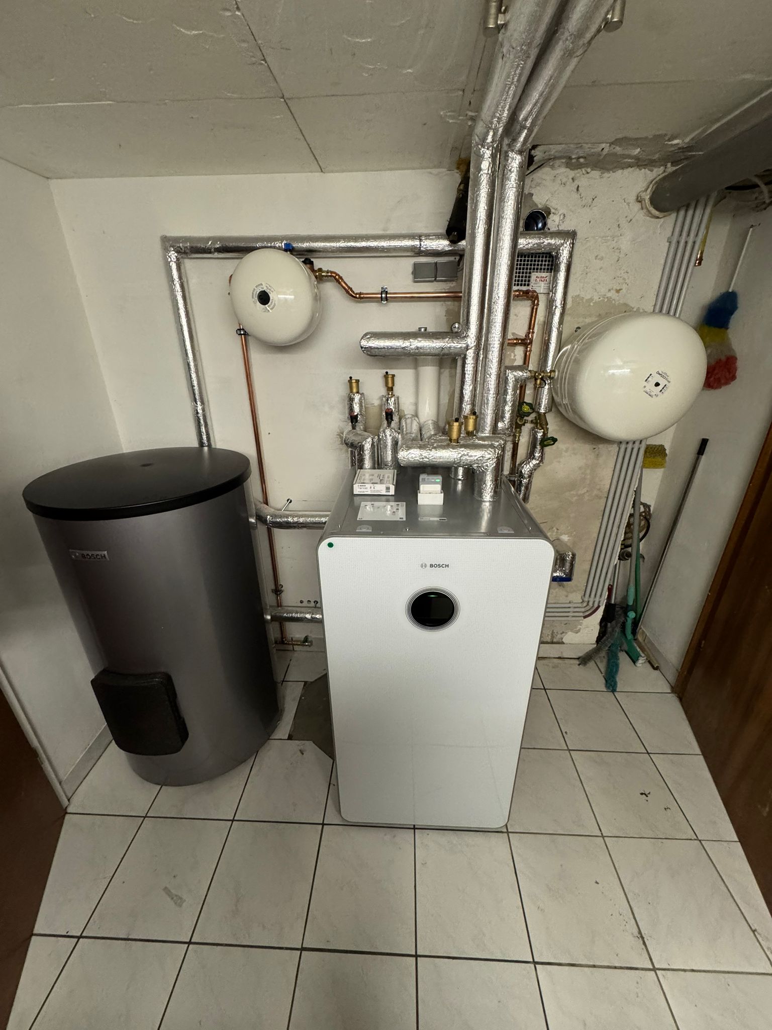 Im Technikraum steht eine Bosch-Wärmepumpe mit integriertem Display, angeschlossen an isolierte Rohrleitungen und einen Pufferspeicher.